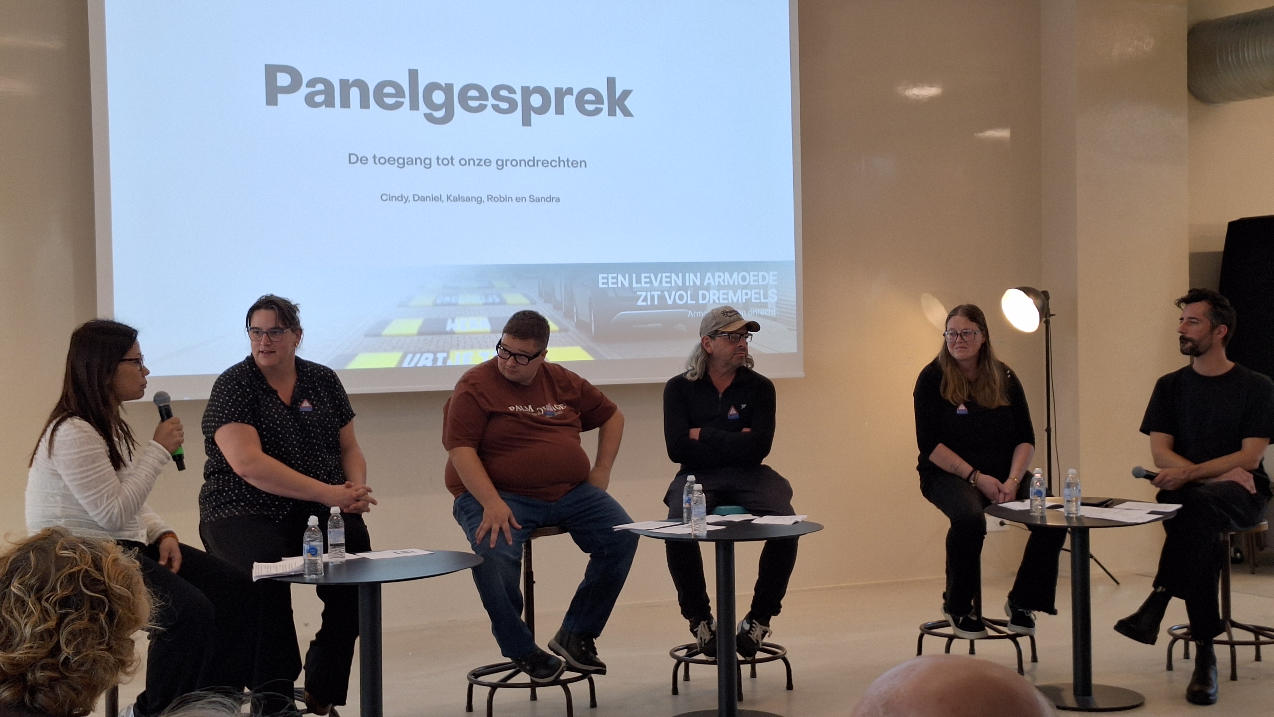 Panelgesprek met 6 mensen. De persoon links heeft een microfoon vast. Ze heeft zwart haar en een witte trui.